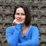 Kvinna med mörkt hår i blå tröja, sittande med handen mot kinden framför en detaljerad, mörk bakgrund.