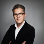 Man med kort mörkt hår och glasögon i svart kavaj, armarna i kors mot en grå bakgrund.