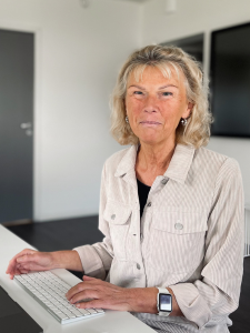 Blond kvinna ler mot kameran medan hon sitter vid ett skrivbord med händerna synligt placerade över ett tangentbord.  tangentbord synligt.