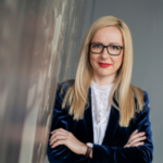 Blond kvinna i glasögon och mörkblå sammetskavaj, poserande mot en reflekterande yta.