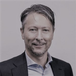  Porträtt av en man med kort mörkt hår, klädd i kostym, leende mot kameran.