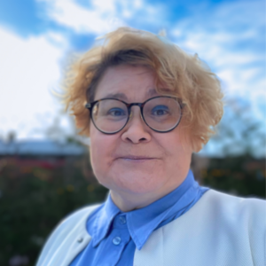 Kortklippt blond kvinna med glasögon, blå blus och vit kofta ler mot kameran.