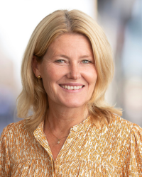Profilbild på kvinna i blont axellångt hår och mönstrad ljusorange och vit blus. Hon ler och kollar in i kameran.