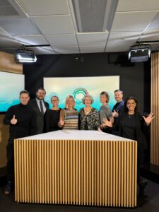 På bild ser man åtta personer som står bakom ett bord. Alla ler mot kameran och viftar på händerna. Bakom syns en skärm med Diversity Charters logga, som är en rund ring med olika färger.