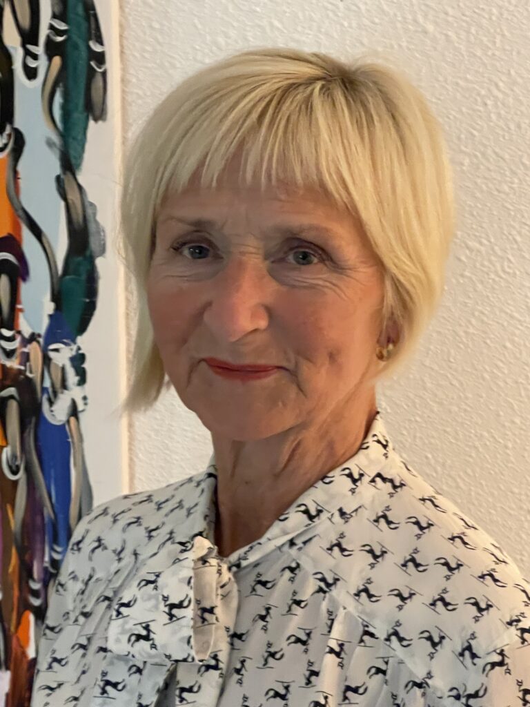 Porträtt på kvinna med ljust kort hår som ler mot kameran. Hon har en vit blus på sig med svart mönster.