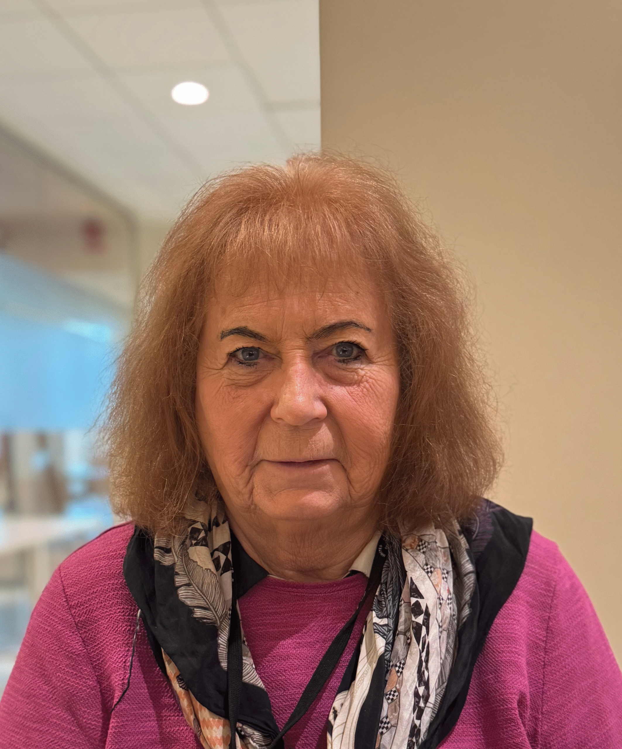 Porträtt av en äldre kvinna med axellångt ljust brunt hår, rosa tröja och mönstrad scarf, inomhus i en ljus miljö.