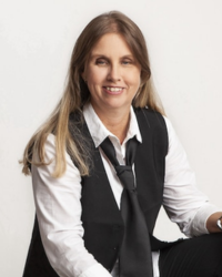 Profilbild på Anna Bergholtz sittandes på en pall i fotostudio. Hon har långt blont hår, klädd i svart väst, vit skjorta, slips, svarta byxor och tittar mot bilden med ett leende
