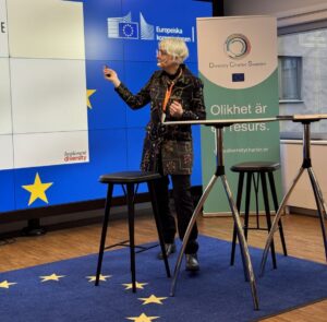 Kvinna mer grått hår pekar på en skärm. På golvet ligger en matta med EUs flagga.