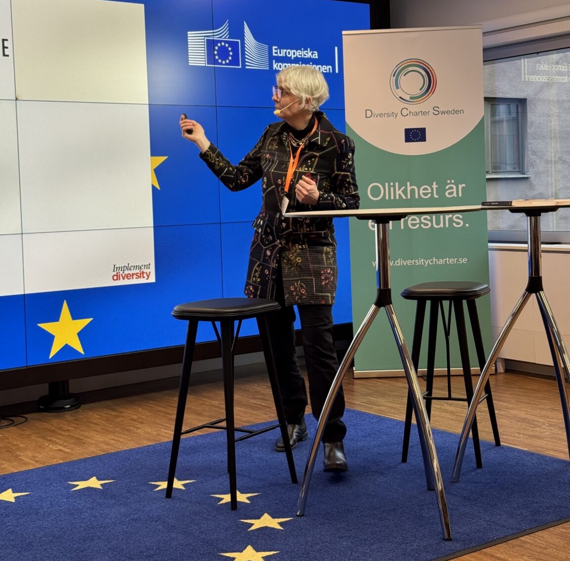 Kvinna mer grått hår pekar på en skärm. På golvet ligger en matta med EUs flagga.