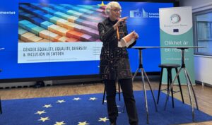 Kvinna med svarta kläder och grått hår håller i ett föredrag.