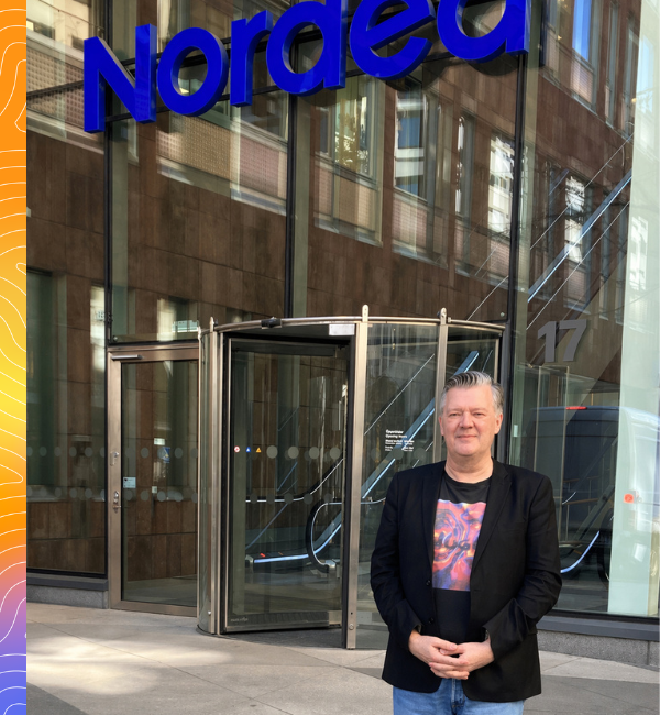 En man med kort grått hår, klädd i svart kostym och blå jeans står utanför Nordeas kontor.