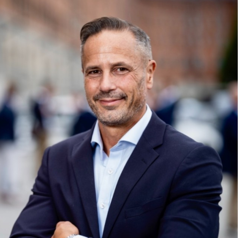 Profilbild på medelåldersman. Gråsbrunt hår. Kollar in i kameran. Mörk kostym.