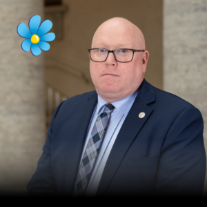 Porträtt på man med flint och glasögin. Iklädd mörkblå kostym. Sverigedemokraternas logotyp. 