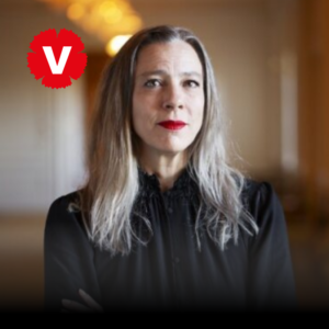 Kvinna i gråblont hår mellanlångt hår. Rött läppstift och svart tröja. Vänsterpartiets logotyp.