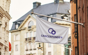 En vit flagga med Cancerfondens blåa logotyp. I bakgrunden en byggnad.