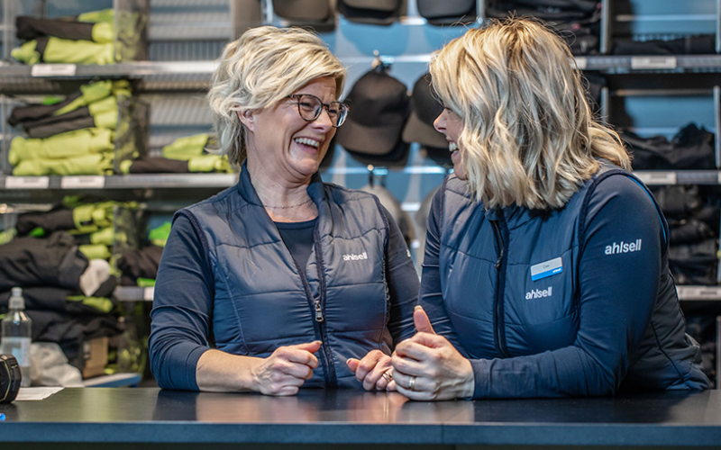 Två kvinnor i medelåldern med blont hår skrattar vända mot varandra, de bär kläder med Ahlsells logotyp. I bakgrunden produkter från Ahlsell.