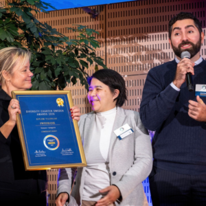 Tre personer på scen under Diversity Charter Sweden Awards 2026. En av dem håller upp ett inramat diplom där Swedbank utses till vinnare i kategorin Långsiktighet, medan en annan talar i mikrofon.