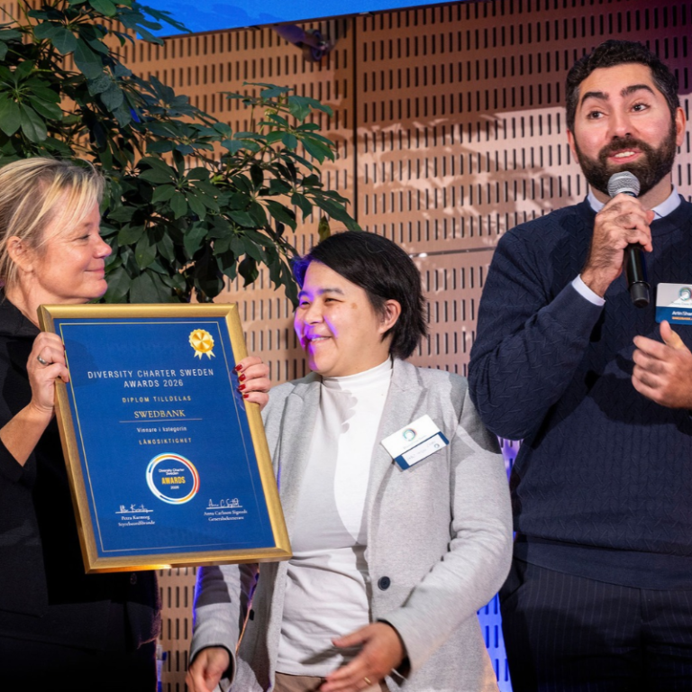 Tre personer på scen under Diversity Charter Sweden Awards 2026. En av dem håller upp ett inramat diplom där Swedbank utses till vinnare i kategorin Långsiktighet, medan en annan talar i mikrofon.