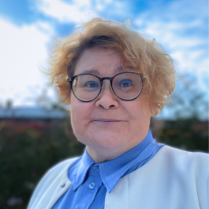 Kortklippt blond kvinna med glasögon, blå blus och vit kofta ler mot kameran.
